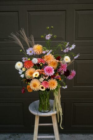 A lush, garden-style bouquet of seasonal dahlias, cosmos, and grasses arranged in a clear glass vase, sitting on a rustic wooden stool in front of a dark garage door.